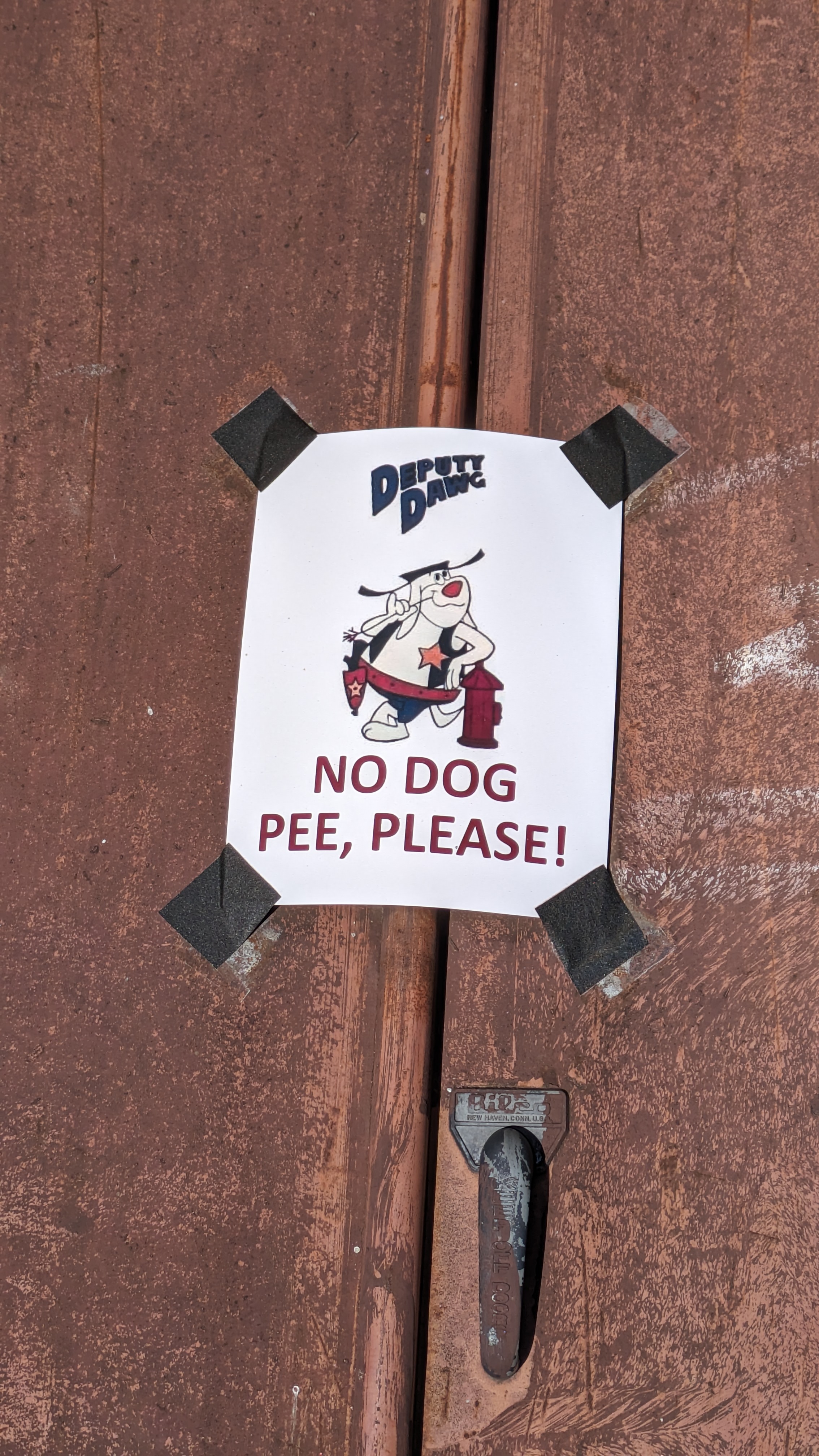 Photograph of a normal letter paper sign duck taped to rusty basement doors. The sign has a cartoon dog labeled Deputy Dawg casually leaning on a fire extinguisher and picking his teeth with a piece of straw. He has a gun in a belt holster and a star badge on his vest. Below him reads NO DOG PEE, PLEASE! in all caps red letters.